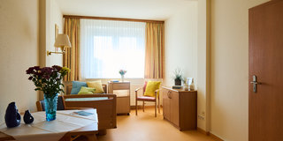 Ein heller Wohnraum mit einem Fenster, durch das Tageslicht strömt. Auf einem Tisch steht eine Vase mit bunten Blumen. Es gibt zwei Stühle und ein Sofa mit bunten Kissen. Die Wände sind hell, und der Boden ist in einem warmen Farbton gehalten.