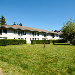 Ein mehrstöckiges Wohngebäude mit einer modernen Fassade steht in einer grünen Umgebung. Der gepflegte Rasen und einige Büsche umgeben das Gebäude. Im Hintergrund sind Bäume zu sehen und der Himmel ist klar und blau.