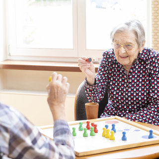 Zwei ältere Personen sitzen sich an einem Tisch gegenüber und spielen ein Brettspiel. Eine Frau mit einer Brille und einem gepunkteten Hemd lächelt und hält einen Spielstein in der Hand, während der andere Spieler sie ansieht. Es gibt bunte Spielsteine auf dem Tisch.