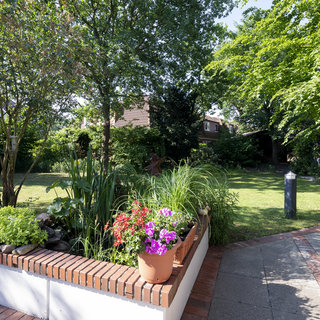 Ein sonniger Garten mit einer bepflanzten Hochbeet umgeben von bunten Blumen in Töpfen. Im Hintergrund sind große Bäume und ein gepflegter Rasen zu sehen. Die angenehme Atmosphäre lädt zum Entspannen im Freien ein.