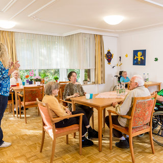 Eine Gruppe älterer Menschen sitzt an Tischen in einem hellen Raum. Sie interagieren miteinander, während eine Person im Vordergrund spricht. An den Wänden hängen dekorative Bilder und bunte Blätter. Fensterlicht und Pflanzen schaffen eine angenehme Atmosphäre.