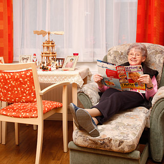 Eine ältere Frau sitzt entspannt in einem bequemen Sessel und liest eine Zeitschrift. Sie trägt bequeme Kleidung und hat graue Haare. Im Hintergrund befindet sich ein Tisch mit dekorativen Gegenständen. Ein warmes, gemütliches Ambiente strahlt aus dem Raum.