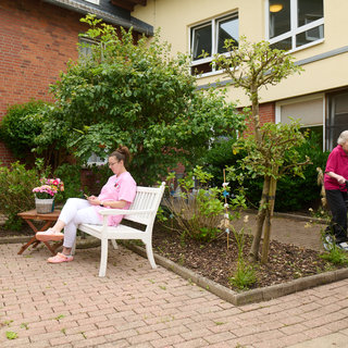 Eine junge Frau sitzt in einem Garten auf einer Bank und schreibt in ein Notizbuch. Eine ältere Dame mit einem Rollator wird von einem Betreuer begleitet. Um sie herum sind Pflanzen und Bäume, die eine angenehme Atmosphäre schaffen.