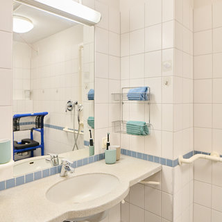 Ein modernes Badezimmer mit weißen Fliesen, einem Spiegel und einem Waschbecken. Neben dem Waschbecken befinden sich Handtücher und Pflegeprodukte. An der Wand ist ein Haltegriff installiert. Im Hintergrund ist ein blauer Stuhl zu sehen.