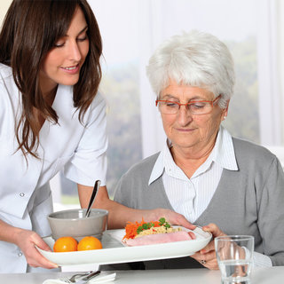 Eine pflegende Person reicht einer älteren Frau einen Teller mit Essen. Die Frau, die grauhaarig ist und eine Brille trägt, schaut aufmerksam auf das Essen, während ein Glas Wasser neben ihr steht.