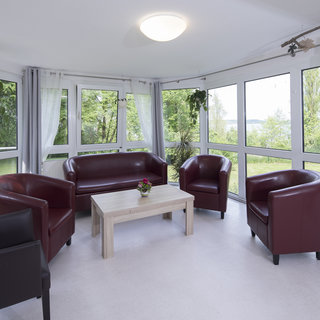 Ein heller, gemütlicher Raum mit großen Fenstern, die Blick auf einen Garten bieten. In der Mitte steht ein kleiner Holztisch, umgeben von vier roten Sesseln und einem schwarzen Sessel. Pflanzen hängen an den Wänden und erhellen die Atmosphäre.