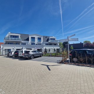 Ein modernes Wohngebäude mit mehreren Etagen steht im Vordergrund. Vor dem Gebäude befinden sich mehrere geparkte Autos. Im Hintergrund ist ein blauer Himmel mit vereinzelten Wolken zu sehen. Neben dem Parkplatz gibt es ein Hinweisschild mit verschiedenen Richtungen.