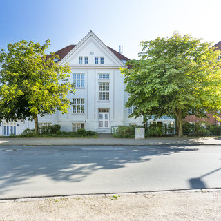 Ein großes, weißes Gebäude mit großen Fenstern und einem roten Dach steht an einer Straße, umgeben von grünen Bäumen und Sträuchern. Rechts im Bild ist ein weiteres, braunes Gebäude sichtbar. Es ist ein heller, sonniger Tag.