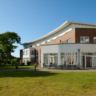 Ein modernes Gebäude mit großen Fenstern und einer einladenden Terrasse steht im Freien. Vor dem Gebäude erstreckt sich ein gepflegter Rasen, umgeben von schattenspendenden Bäumen. Der Himmel ist klar und blau, was eine freundliche Atmosphäre schafft.