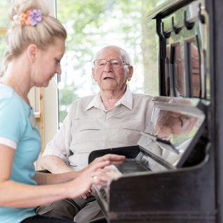 Eine Frau spielt Klavier, während ein älterer Mann aufmerksam zusieht. Die Szene vermittelt eine Atmosphäre der Freude und des Miteinanders, möglicherweise in einem entspannten, hellen Raum.