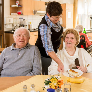 Eine ältere Frau sitzt an einem Tisch und lächelt, während eine Mitarbeiterin ihr ein Dessert reicht. Neben ihr sitzt ein älterer Mann, der ebenfalls lächelt. Der Tisch ist gedeckt mit einem Blumenarrangement und Getränken. Der Raum hat eine helle, freundliche Atmosphäre.