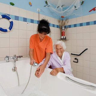 Eine Pflegekraft hilft einer älteren Frau beim Baden. Die Frau, in einem lilafarbenen Bademantel, lächelt während die Pflegekraft den Wasserhahn bedient. Die Umgebung ist hell und freundlich gestaltet, mit maritimen Dekorationselementen an den Wänden.
