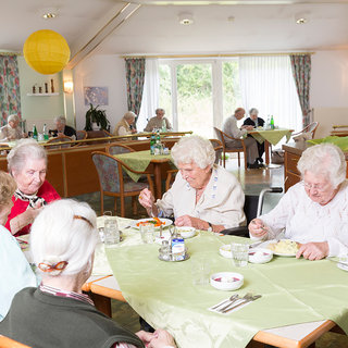 Eine Gruppe älterer Frauen sitzt an einem Tisch in einem hellen Raum mit großen Fenstern. Sie essen gemeinsam eine Mahlzeit, während weitere Bewohner im Hintergrund ebenfalls an anderen Tischen speisen. Der Tisch ist mit grüner Tischdecke und Gläsern gedeckt.