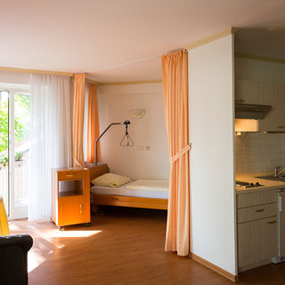 Der Bildinhalt zeigt ein helles Zimmer mit einem Bett, einem Tisch und einer Küchenzeile. Große Fenster lassen Tageslicht herein, und orangefarbene Vorhänge rahmen den Raum ein. Eine Stehlampe steht neben dem Bett, und der Boden ist aus Holz.