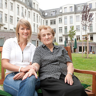 Eine junge Frau sitzt neben einer älteren Frau auf einer Bank im Freien. Beide lächeln und wirken freundlich. Im Hintergrund sind Gebäude und einige Bäume sichtbar. Die Szene strahlt Wärme und Verbundenheit aus.