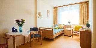 Das Bild zeigt ein helles Zimmer mit einem Einzelbett, einem Tisch mit Stühlen und einer Fensterbank, die mit Pflanzen und Kissen dekoriert ist. An der Wand hängt ein Bild. Der Raum wirkt einladend und freundlich, mit einem warmen Farbton.