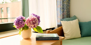 Auf einem Holztisch steht eine Vase mit lila und rosa Hortensien. Daneben liegt ein Buch mit dem Titel "In Stadtlichtersker Nacht". Im Hintergrund sind ein Fenster mit Vorhängen und eine Couch mit Kissen sichtbar. Das Licht fällt sanft in den Raum.