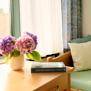 Auf einem Holztisch steht eine Vase mit lila und rosa Hortensien. Daneben liegt ein Buch mit dem Titel "In Stadtlichtersker Nacht". Im Hintergrund sind ein Fenster mit Vorhängen und eine Couch mit Kissen sichtbar. Das Licht fällt sanft in den Raum.