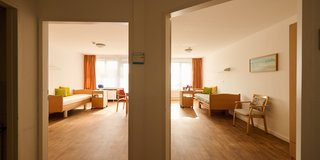 Ein heller Wohnraum mit zwei angrenzenden Bereichen. Auf der linken Seite ist ein gemütliches Sofa und ein Tisch, auf der rechten Seite stehen weitere Betten und Stühle. Große Fenster sorgen für viel Tageslicht. Die Wände sind in neutralen Farben gehalten.