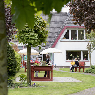 Ein gepflegter Garten mit Rasenflächen, Bäumen und Blumen. In der Mitte stehen rote Tisch-Bänke sowie Sonnenschirme. Zwei Personen unterhalten sich, während andere auf Bänken sitzen. Im Hintergrund ist ein weißes Gebäude mit Fenstern und bunten Dekorationen zu sehen.