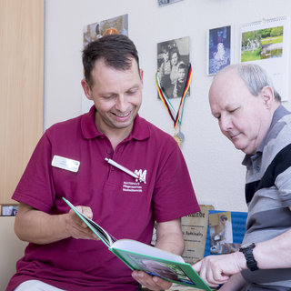 Ein Mitarbeiter setzt sich mit einem älteren Mann zusammen und zeigt ihm ein Buch. Beide lächeln, während sie auf die Seiten schauen. Der Raum ist freundlich gestaltet und mit Erinnerungsfotos dekoriert.