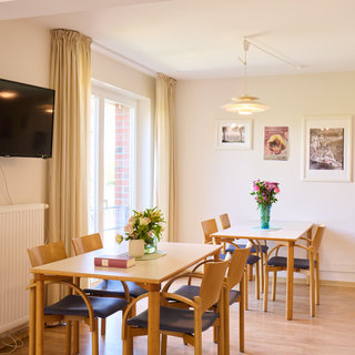 Ein helles Zimmer mit drei Tischen und Stühlen, dekoriert mit Blumen. An der Wand hängen einige Bilder. Ein Fernseher ist an der Wand befestigt, und ein Heizkörper ist sichtbar. Fenster lassen Tageslicht herein.