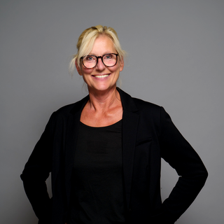 Eine lächelnde Frau mit blonden Haaren trägt eine schwarze Bluse und einen schwarzen Blazer. Sie steht vor einem grauen Hintergrund und hält eine Hand an ihrer Hüfte. Die Brille mit runden Gläsern unterstreicht ihren freundlichen Ausdruck.