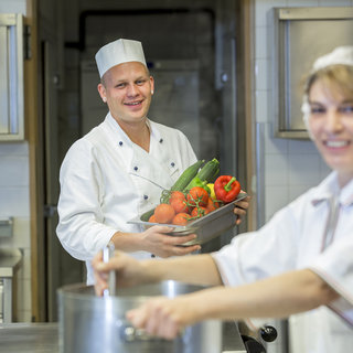 Ein Koch lächelt, während er eine Schale mit frischem Gemüse in der Hand hält. Im Hintergrund bereitet eine Kollegin in einer weißen Schürze und Haube eine Suppe zu. Die Szene spielt in einer modernen Küche.