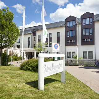 Das Bild zeigt das Gebäude der "Barbara-Ulmann-Stift". Im Vordergrund steht ein weißes Schild mit dem Namen der Einrichtung. Um das Gebäude gibt es Rasenflächen und einige Bäume. Im Hintergrund sind mehrere Fenster und Flaggen sichtbar.