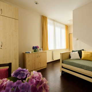 Ein freundliches Zimmer mit einem Bett, einem großen Fenster, das Tageslicht hereinlässt, und hellen Vorhängen. An der Wand steht ein Schrank und ein Tisch mit einer blühenden Pflanze. Das Gesamtbild vermittelt eine gemütliche und einladende Atmosphäre.