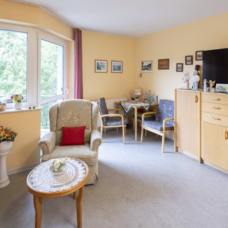 Das Bild zeigt ein helles Wohnzimmer mit einem Fenster, das Blick ins Freie bietet. In der Ecke steht ein bequemer Sessel mit einem roten Kissen. Ein Couchtisch mit einer Tischdecke ist ebenfalls sichtbar, und die Wände sind mit Bildern dekoriert. Ein TV-Schrank rundet die Einrichtung ab.