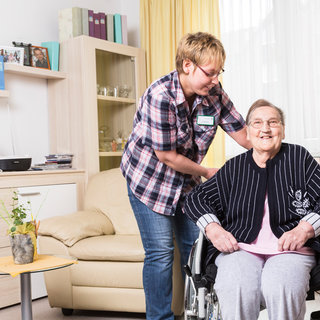 Eine ältere Frau sitzt in einem Rollstuhl und lächelt. Eine Pflegerin steht neben ihr und spricht freundlich mit ihr. Der Raum ist hell und gemütlich eingerichtet, mit einem Sofa und Pflanzen im Hintergrund.