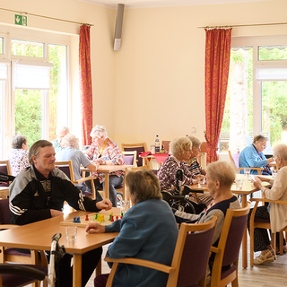 Eine gemütliche Cafeteria mit mehreren älteren Menschen, die an Tischen sitzen und sich unterhalten. Einige spielen mit einem Spielmaterial. Große Fenster sorgen für viel Tageslicht und der Raum ist freundlich eingerichtet.