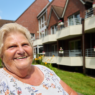 Eine lächelnde ältere Frau mit blonden Haaren sitzt im Freien vor einem Mehrfamilienhaus. Der Hintergrund zeigt Balkone mit Blumenkästen und einen blauen Himmel. Die Atmosphäre wirkt freundlich und einladend.