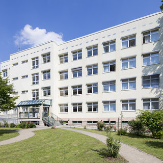 Ein modernes, mehrstöckiges Gebäude in hellem Beige steht im Mittelpunkt. Es hat große Fenster, die Tageslicht einlassen. Vor dem Gebäude erstreckt sich ein gepflegter Garten mit Wegen, Bäumen und Sträuchern, der eine einladende Atmosphäre schafft.