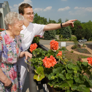 Eine ältere Frau und ein Pfleger stehen auf einem Balkon und schauen zusammen auf einen Garten. Der Pfleger zeigt mit dem Finger auf etwas im Freien, während die Frau aufmerksam lauscht. Bunte Blumen, hauptsächlich Geranien, sind im Vordergrund sichtbar.