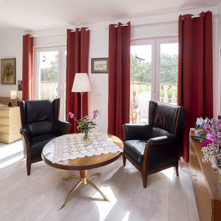 Ein helles Zimmer mit einem Bett, zwei schwarzen Sessel, einem runden Holztisch mit Tischdecke und einer Blumenvase. Rote Vorhänge umrahmen große Fenster, die auf einen Balkon blicken. An den Wänden sind Bilder und Fotos aufgehängt. Der Boden ist aus hellem Holz.