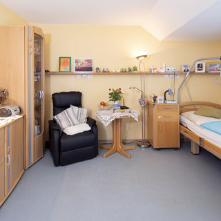 Das Bild zeigt ein gemütliches Schlafzimmer mit gelben Wänden. Auf der linken Seite befindet sich ein Kommode und ein Sessel. In der Mitte steht ein kleiner Tisch mit einer Tischlampe und einer Blumenvase. Rechts ist ein Pflegebett mit einer Decke und Haltegriffen.