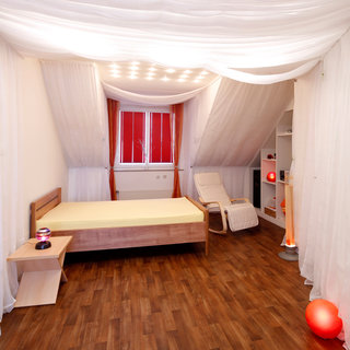 Das Bild zeigt ein modernes Schlafzimmer mit einem Holzbettrahmen, einem Nachttisch sowie einem bequemen Sessel. Die Wände sind mit weißen Vorhängen dekoriert, und es gibt eine Warmlichtlampe sowie eine rote Lampe in der Ecke. Die Atmosphäre wirkt hell und einladend.