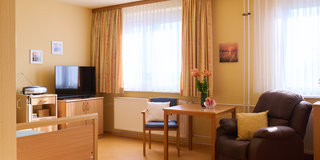 Ein helles Zimmer mit großen Fenstern, durch die Licht strömt. Ein Holzregal mit Fernseher, ein Tisch und zwei Stühle sind sichtbar. In der Ecke steht ein bequemer Sessel. Blumen in einer Vase auf dem Tisch bringen Farbe in den Raum. Die Wände sind in warmen Farbtönen gehalten.