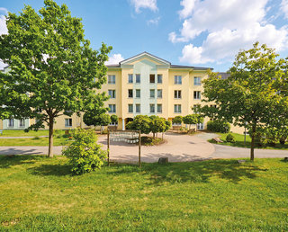 Ein hellgelbes Gebäude mit mehreren Fenstern steht in einem grünen Bereich, umgeben von großen Bäumen und einer gepflegten Rasenfläche. Am Vordergrund gibt es einen gepflasterten Weg und einige Bänke. Der Himmel ist blau mit einigen Wolken.