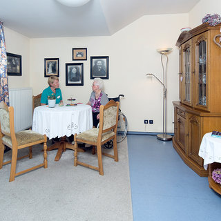 Eine junge Pflegekraft und eine ältere Dame sitzen an einem kleinen Tisch in einem hellen Raum. Der Tisch ist mit einer Tischdecke und einer Pflanze dekoriert. Im Hintergrund sind gerahmte Bilder an der Wand und ein Schrank mit Gläsern zu sehen.