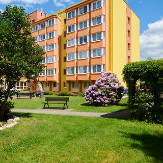 Ein mehrstöckiges, gelbes Wohngebäude umgeben von einem gepflegten Garten. Im Vordergrund stehen zwei Bänke, und zu sehen sind Blumen und grüne Sträucher. Der Himmel ist blau und mit einigen Wolken versehen.