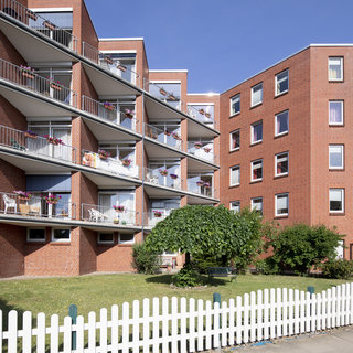 Ein mehrstöckiges Wohngebäude aus rotem Ziegelstein mit Balkonen, die mit Blumenkästen dekoriert sind. Im Vordergrund befindet sich ein kleiner Garten mit einem weißen Zaun. Die Umgebung ist hell und sonnig, was eine einladende Atmosphäre schafft.