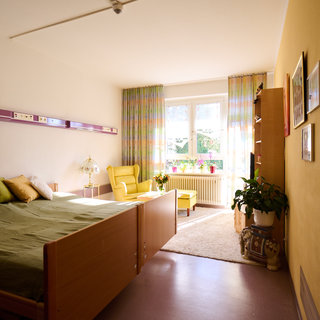 Ein helles Zimmer mit einem Bett, Kissen und einem gemütlichen Sessel. Große Fenster mit bunten Vorhängen lassen viel Licht herein. Ein kleiner Tisch mit einer Lampe und eine Pflanze verleihen dem Raum eine einladende Atmosphäre. 