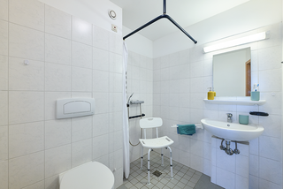 Ein barrierefreies Badezimmer mit hellen Fliesen. Es gibt eine Dusche mit Haltegriffen, einen Duschstuhl, ein Waschbecken mit Handtuch und zwei Seifenspendern sowie eine Toilette. Ein Spiegel und eine Deckenleuchte sorgen für ausreichende Beleuchtung.