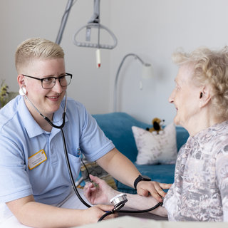 Eine Pflegekraft mit Kurzhaarschnitt und Brille führt eine Untersuchung bei einer älteren Frau durch. Die Pflegekraft betrachtet lächelnd das digitale Messgerät, während die Frau entspannt und freundlich schaut. Die Umgebung wirkt hell und freundlich, mit einem modernen, gemütlichen Wohnraum im Hintergrund.