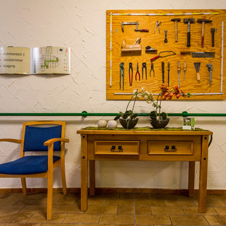 Im Bild ist ein Tisch mit zwei Schubladen und zwei Blumentöpfen darauf zu sehen. An der Wand hinter dem Tisch hängen verschiedene Werkzeuge, darunter Hammer, Zangen und Schraubenschlüssel, die ordentlich angeordnet sind. Neben dem Tisch steht ein blauer Stuhl.