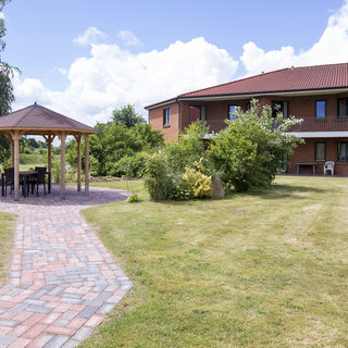 Eine gepflasterte Weg führt zu einem rundem Pavillon mit Sitzmöglichkeiten. Im Hintergrund befindet sich ein zweigeschossiges Gebäude mit Balkonen. Der Garten ist gepflegt, mit Rasen und Sträuchern, unter einem klaren Himmel.