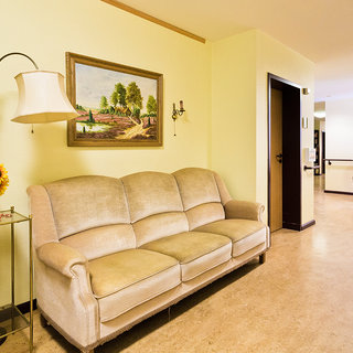 Ein gemütliches Wohnzimmer mit einem hellen, beigen Sofa. An der Wand hängt ein Landschaftsgemälde. Neben dem Sofa steht eine Lampe und ein Beistelltisch mit Sonnenblumen. Im Hintergrund führt eine Tür zu einem weiteren Raum. Der Boden ist hell und wirkt einladend.